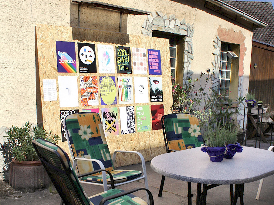 Aufnahme im Hof des Hauses. Im Vordergrund Gartentisch- und stühle, dahinter eine Plakatwand mit Miniaturen der Plakate im Innern.