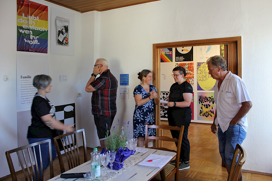 Aufnahme des ersten Ausstellungsraums mit einigen Plakaten, dem Tisch für die Diskussion im Vordergrund, sowie einigen Gästen. Im Hintergrund der zweite Raum der Ausstellung.